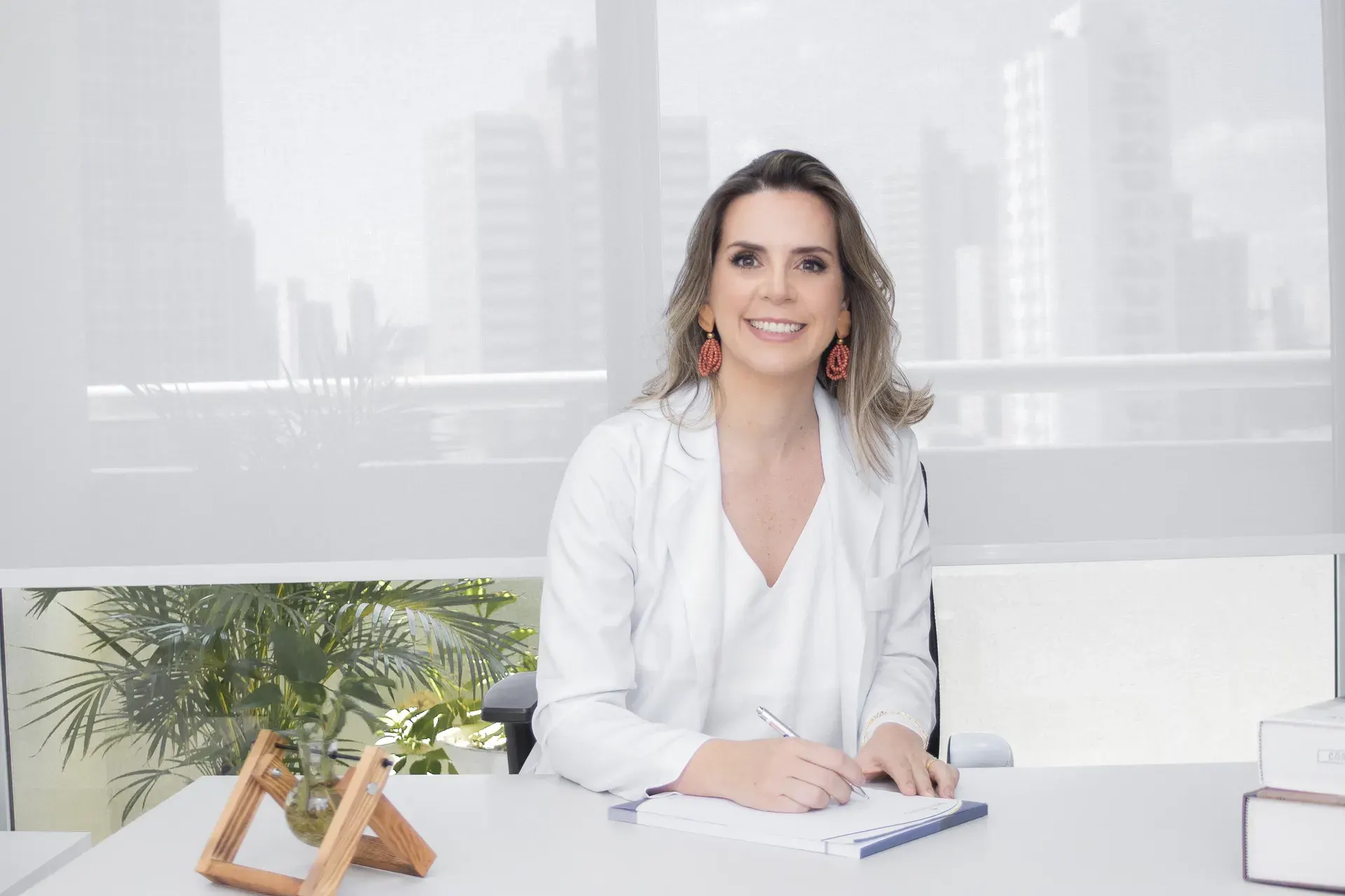 Dra. Aline De Bortoli de jaleco, sentada à mesa do consultório